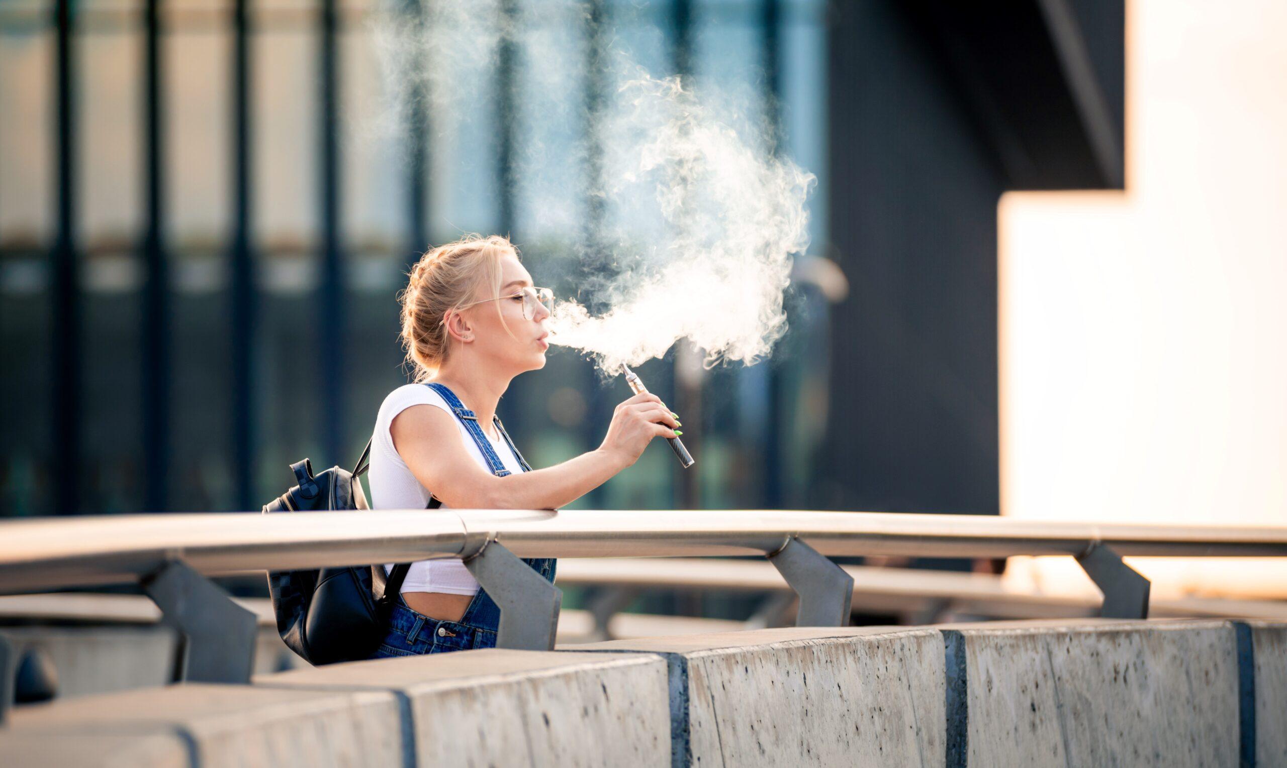 Eine Frau steht draußen auf einer Brücke und dampft eine Einweg-E-Zigarette, während sie eine große Rauchwolke ausatmet. Sie trägt eine Brille und hat einen Rucksack dabei.
