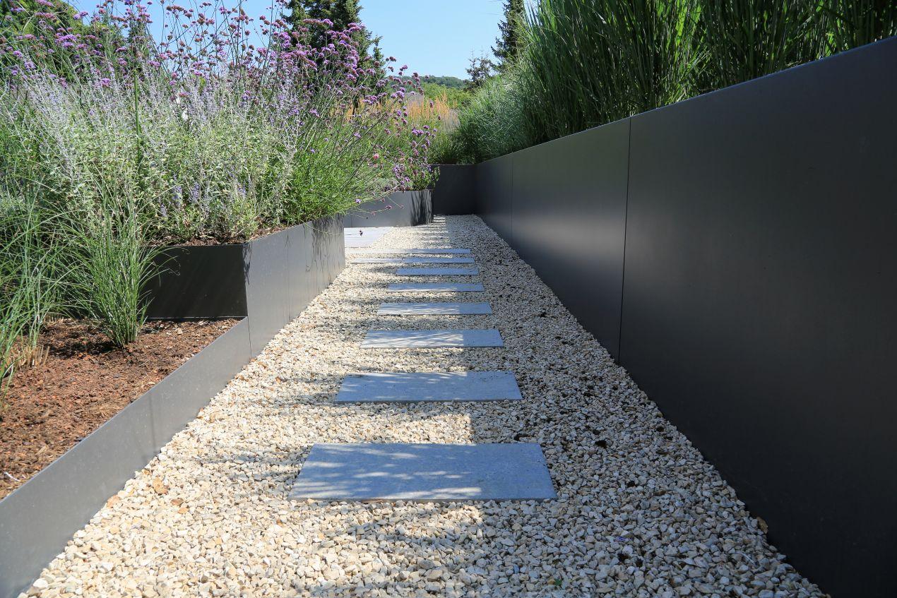Moderner Gartenweg mit grauen Trittplatten auf hellem Zierkies, eingerahmt von Stahlkanten und üppiger Bepflanzung.
